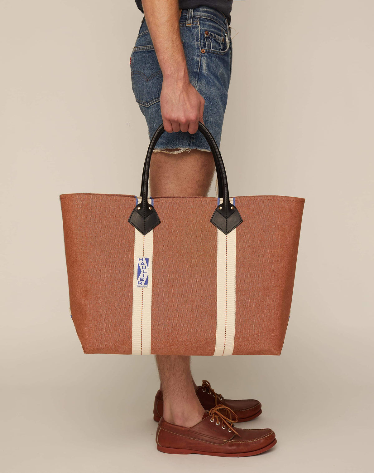 Image of person holding classic canvas tote bag in tan colour with black leather handles and contrasting natural ecru stripes.