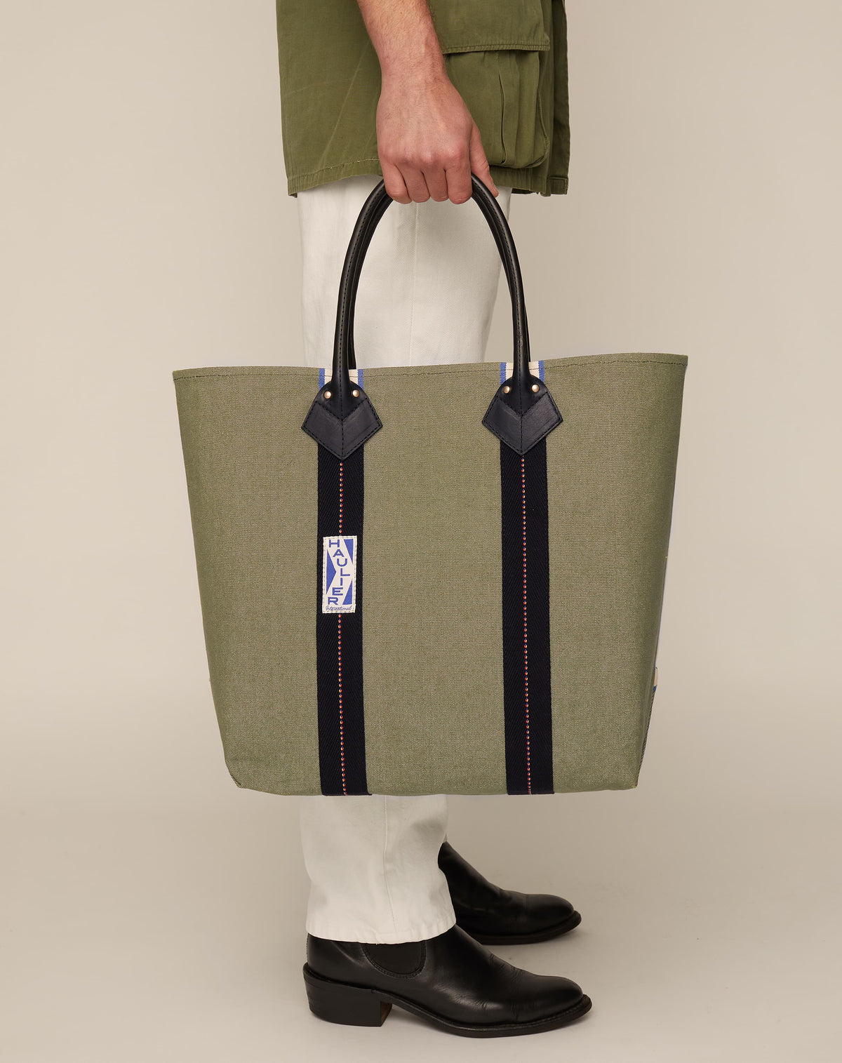 Image of person in black boots and white trousers holding medium-sized classic canvas tote bag in sage colour with black leather handles and contrasting stripes.