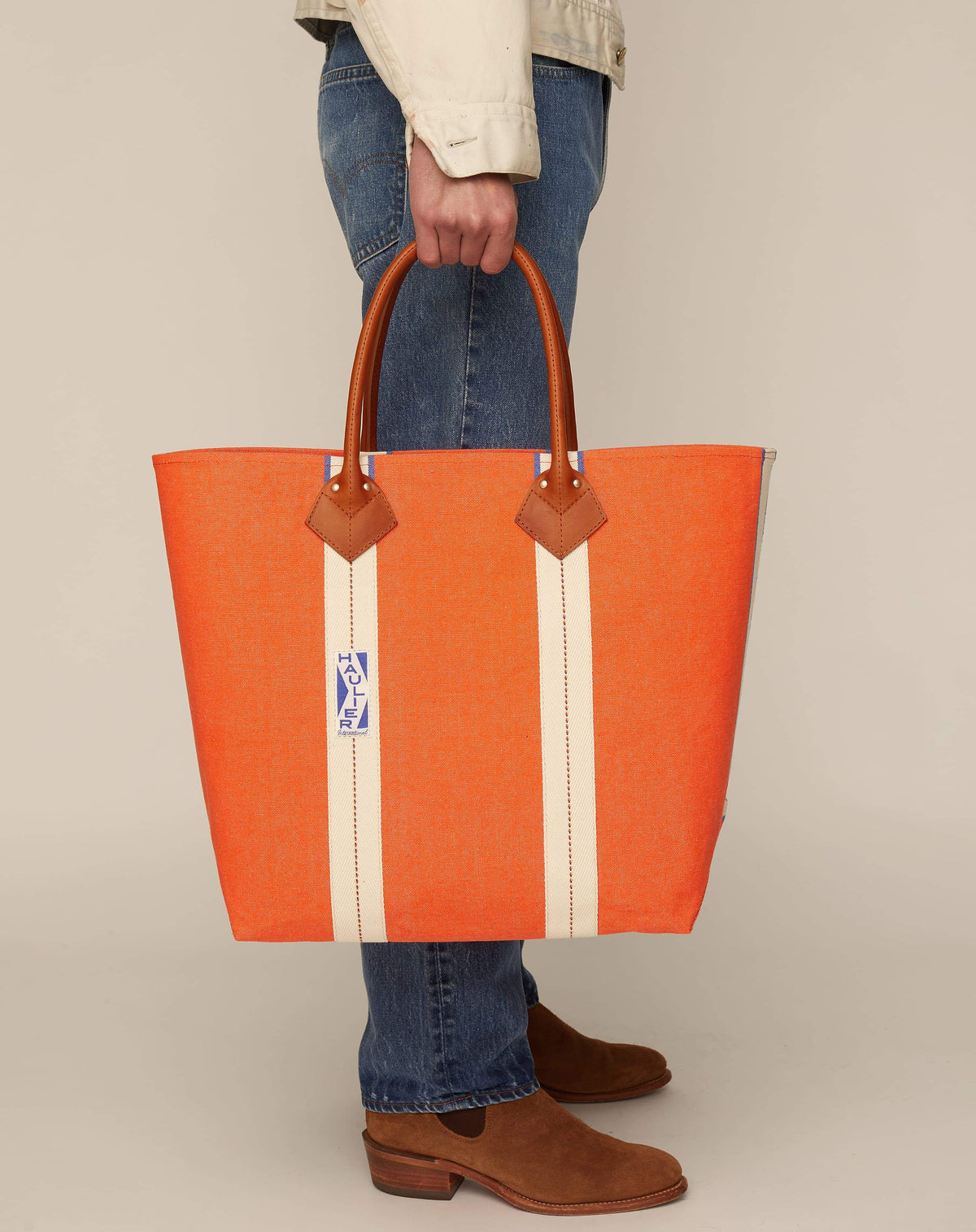 Image of person in jeans holding medium-sized classic canvas tote bag in orange colour with tan leather handles and contrasting stripes.