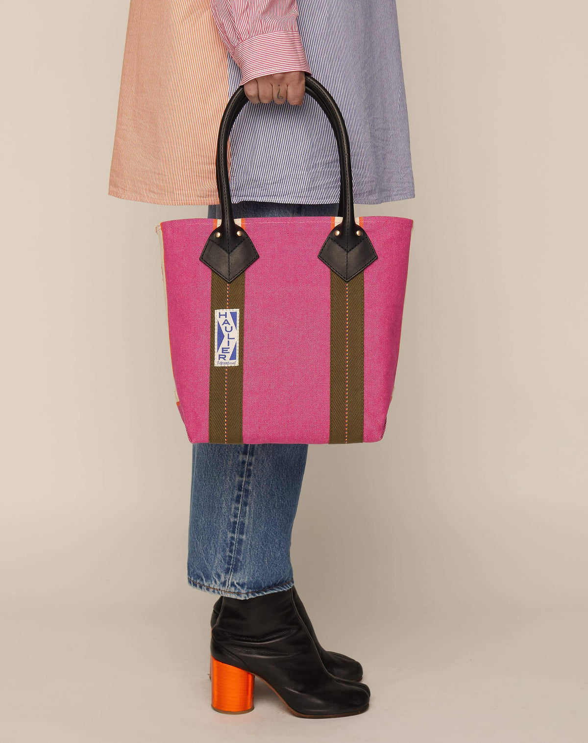 Image of person in jeans, boots and striped shirt holding a small classic canvas tote bag in fuchsia colour with black leather handles and contrasting stripes.