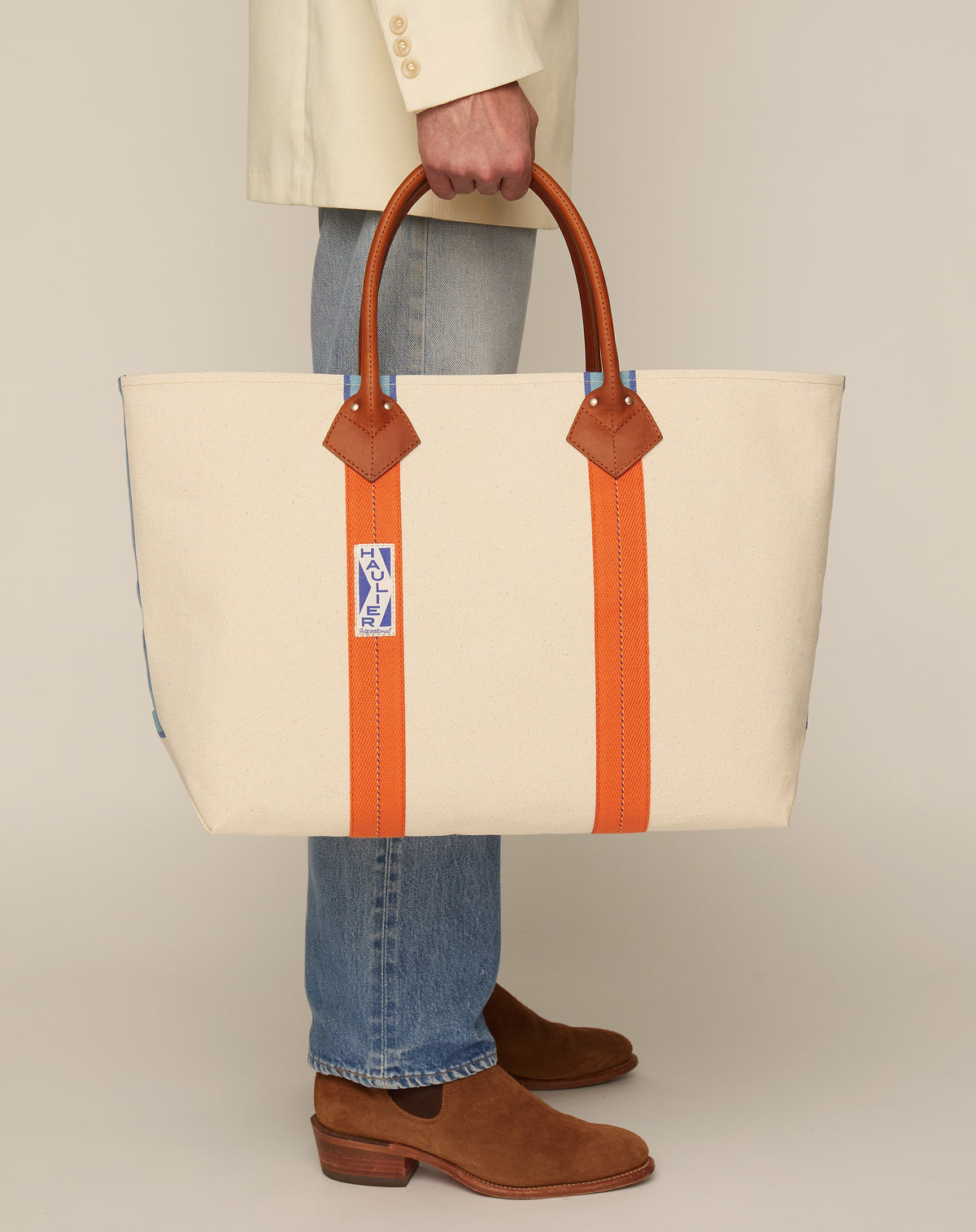 Image of man holding a classic canvas tote bag in natural ecru colour with leather handles and contrasting orange stripes.