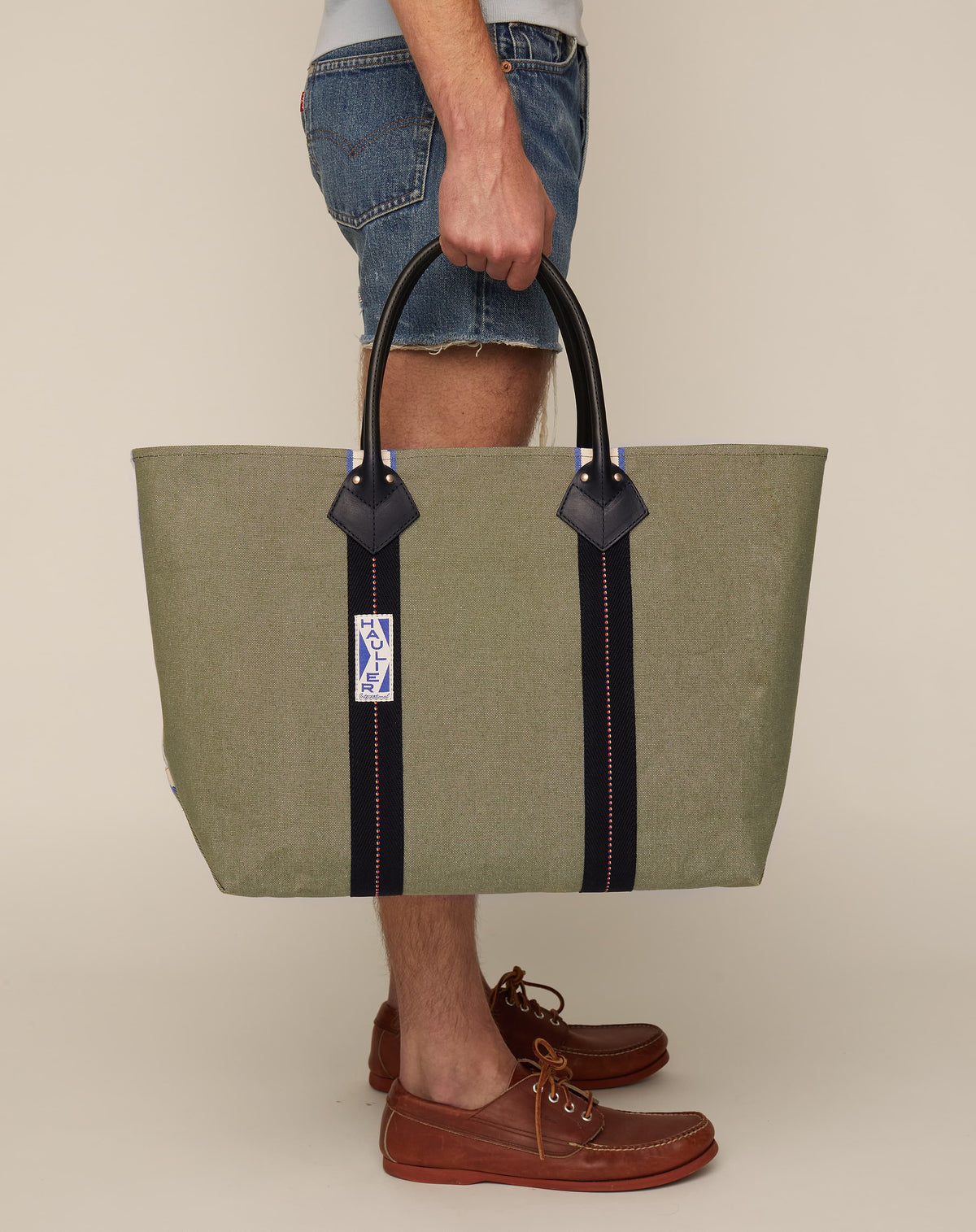 Image of person holding a classic canvas tote bag in sage colour with black leather handles and contrasting black stripes.