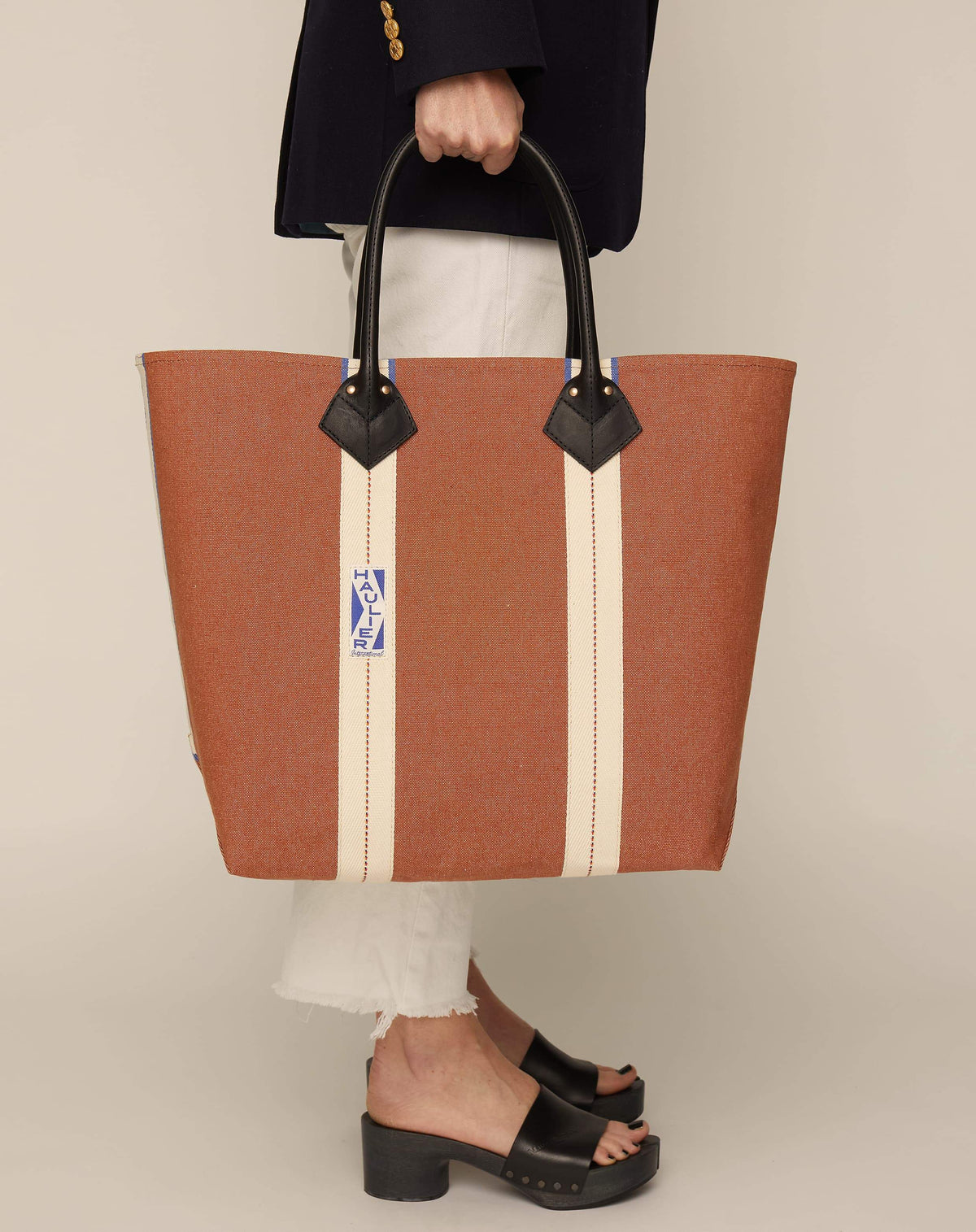 Image of person in white trousers and black slides holding medium-sized classic canvas tote bag in tan colour with black leather handles and contrasting stripes.