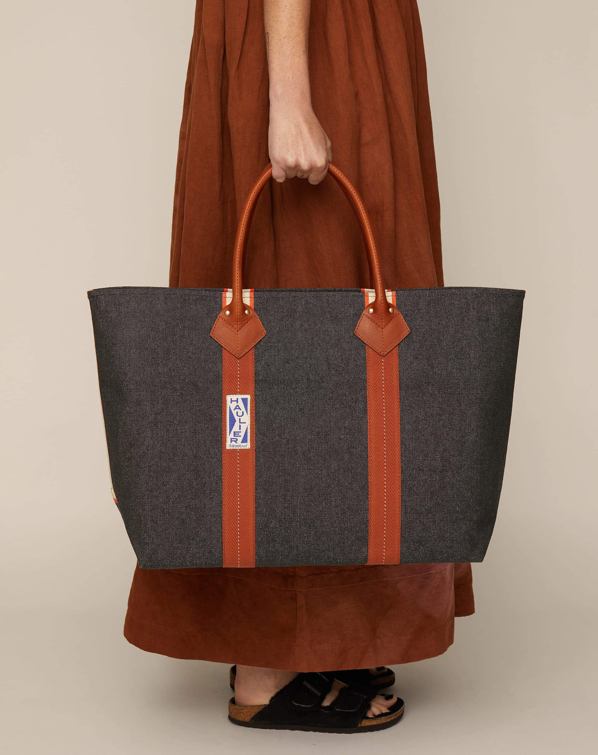 Image of person in brown skirt holding a classic canvas tote bag in washed black colour with tan leather handles and contrasting stripes.