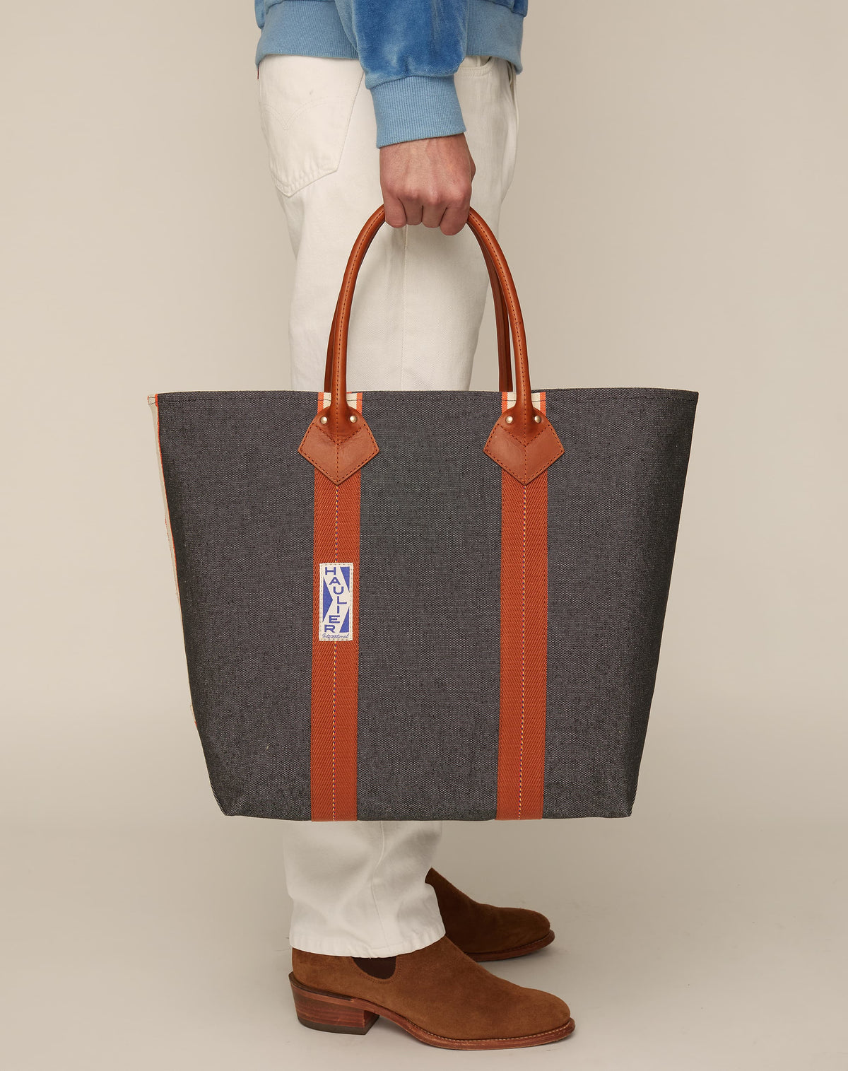 Image of person in white trousers and tan boots holding a medium-sized classic canvas tote bag in washed black colour with tan leather handles and contrasting stripes.