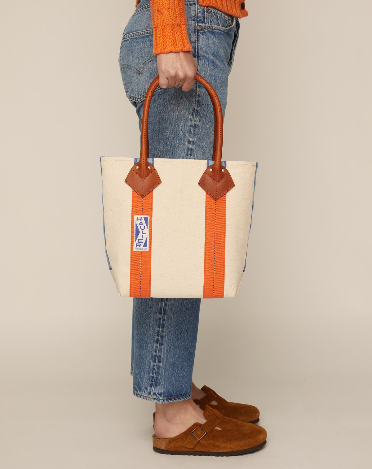 Image of person in blue jeans and tan slides holding a small classic canvas tote bag in natural ecru colour with tan leather handles and contrasting stripes.