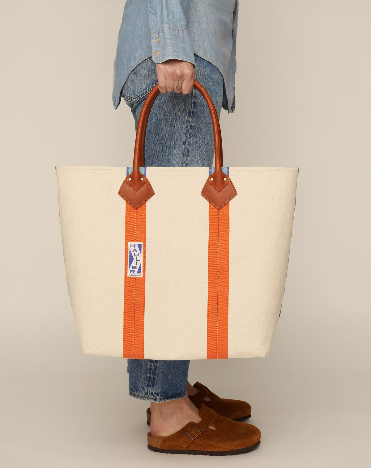 Image of person in blue jeans holding a medium-sized classic canvas tote bag in natural ecru colour with tan leather handles and contrasting stripes.