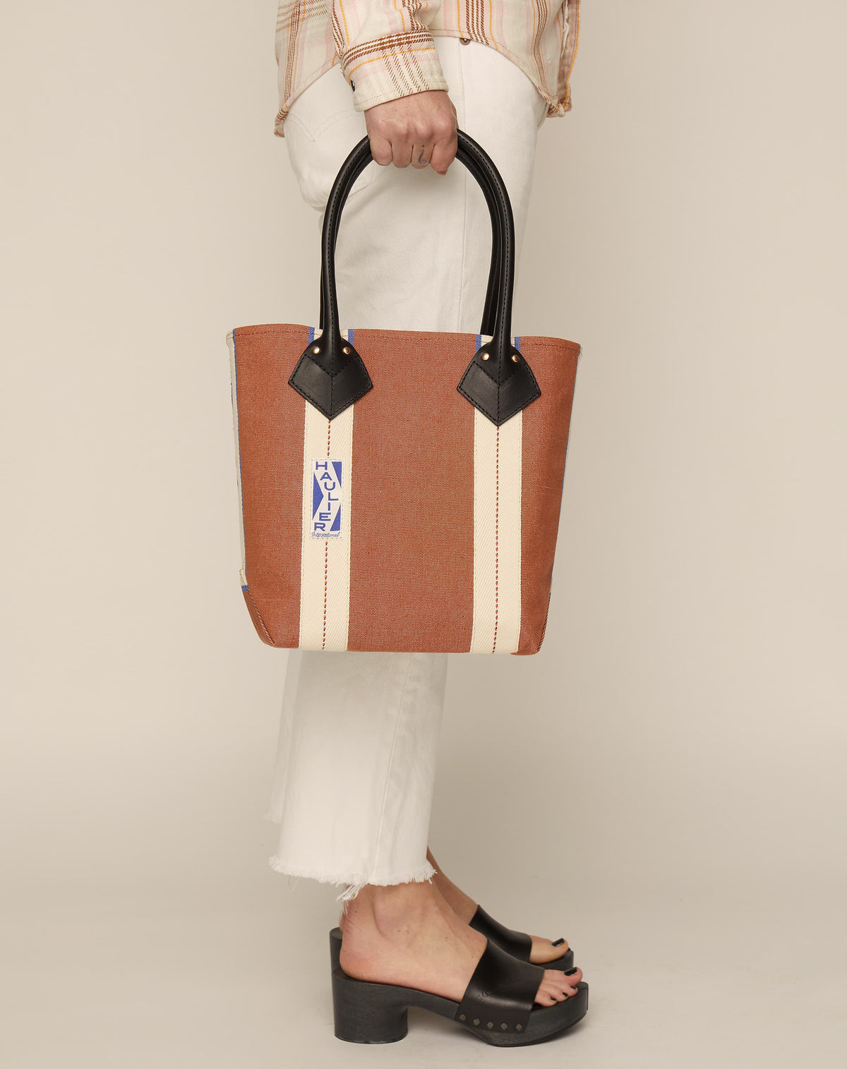 Image of person in white jeans and black slides holding a small classic canvas tote bag in tan colour with black leather handles and contrasting stripes.