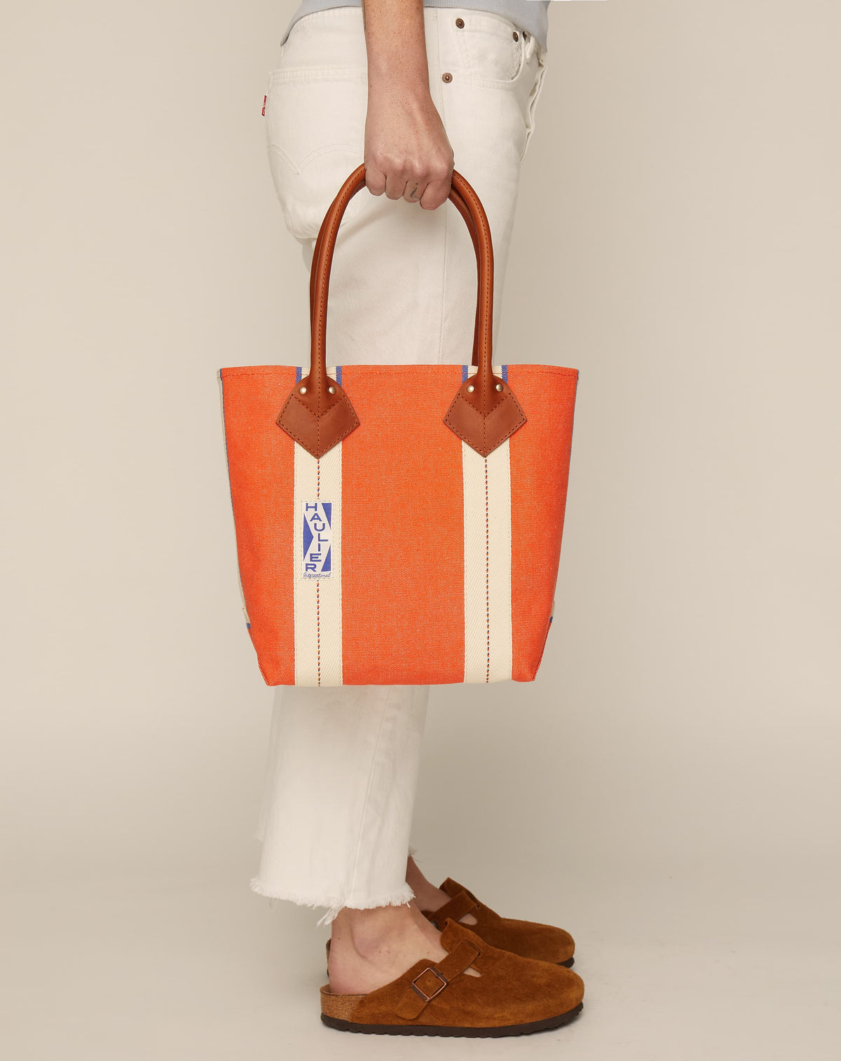 Image of person in white jeans and tan slides holding a small classic canvas tote bag in orange colour with tan leather handles and contrasting stripes.