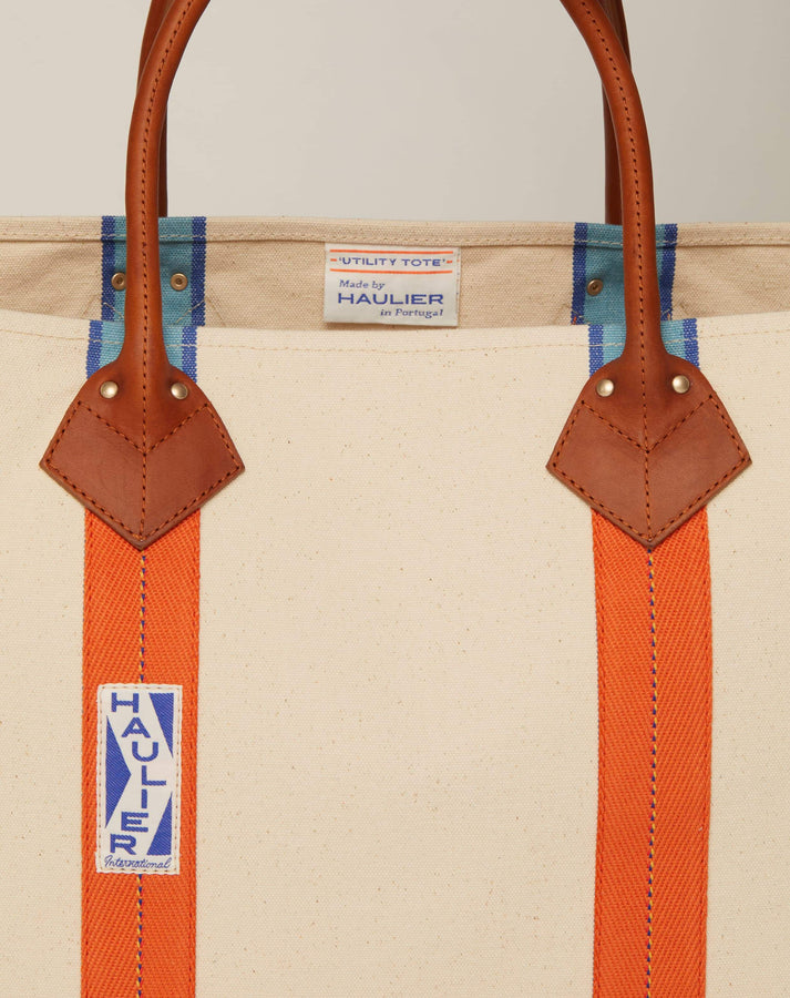 Close-up cropped image of classic canvas tote bag in natural ecru colour with leather handles and contrasting orange stripes and HAULIER logo.