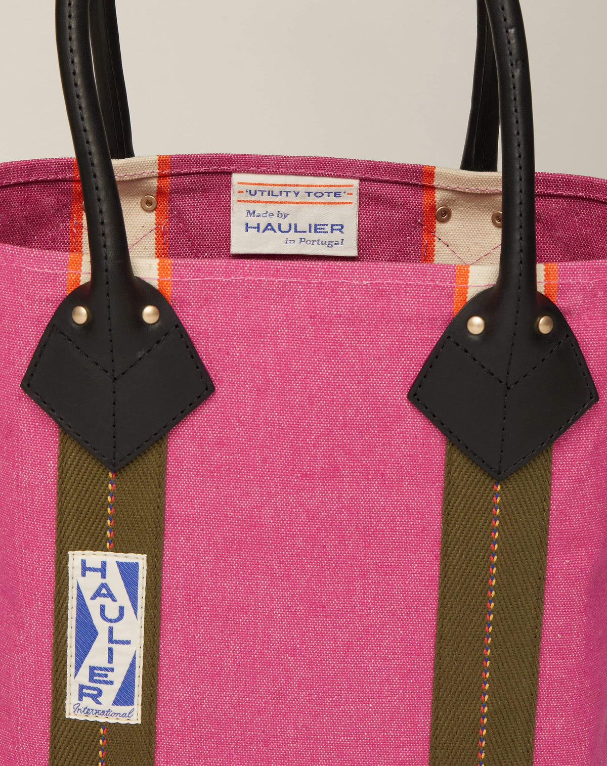 Close-up image of small classic canvas tote bag in fuchsia colour with black leather handles and contrasting stripes and HAULIER branding.